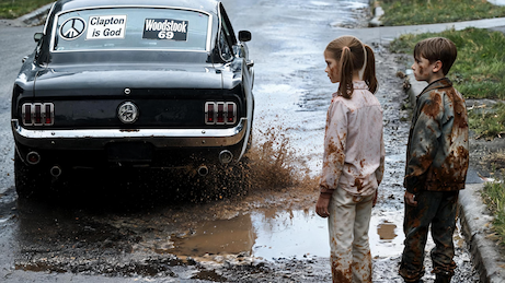A Vintage Mustang drives past two children on a wet road and splashes them with mud from a puddle.  On the back window of the car are stickers, such as 'Clapton is God' and 'Woodstock 69'